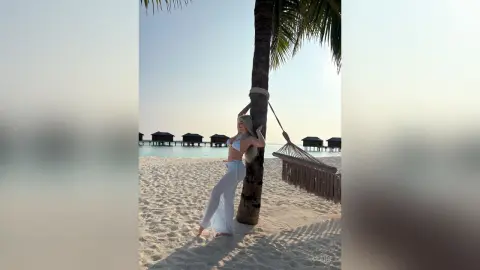 Blonde woman in a white bikini and sheer cover-up leaning against a palm tree on a sunny tropical beach.