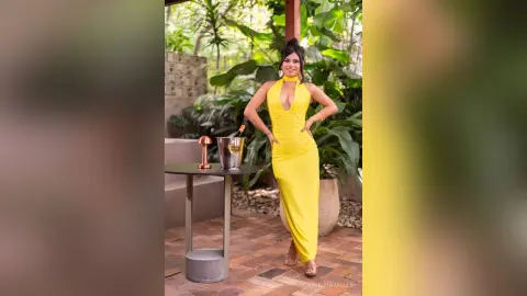 Smiling woman in a bright yellow halter maxi dress standing by a patio table in a lush tropical garden.