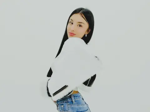 Ema_Velvet poses against a white background wearing a white furry jacket and blue denim jeans, looking over her shoulder with long black hair.