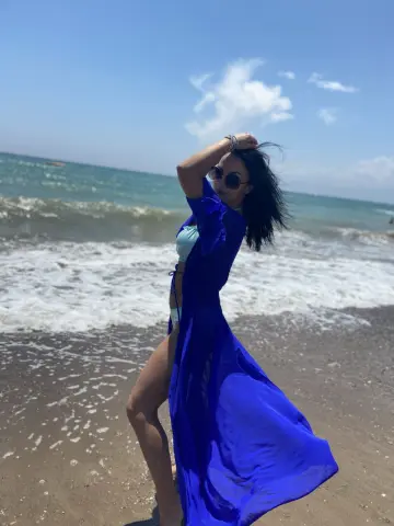 Issadorra poses on a sandy beach wearing a blue open robe and light blue bikini, looking over her shoulder with her hand in her dark hair.