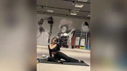 NicoleBella sits on a black yoga mat in a gym, wearing a black crop top and black leggings.