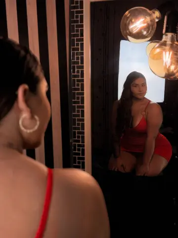 Young woman in a red dress looking into a mirror in a moody, dimly lit room with warm glowing light bulbs.