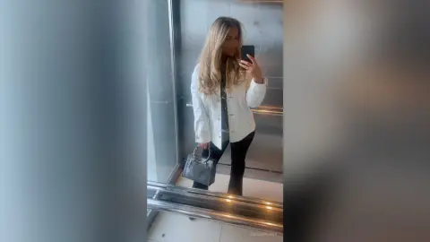 Blonde woman in a white jacket and black pants holding a grey handbag in a modern hallway mirror selfie.