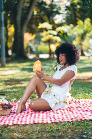 Smiling Black woman in a white dress enjoying a sunny garden picnic on a red checkered blanket.