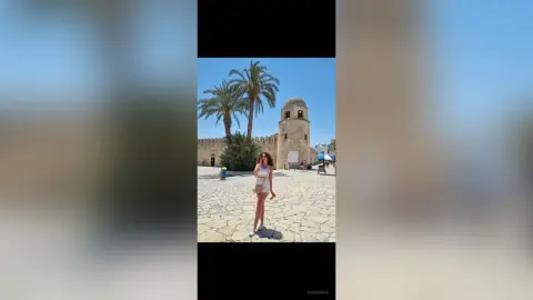 Young woman in a white summer outfit standing in a sunny historic stone courtyard with palm trees.