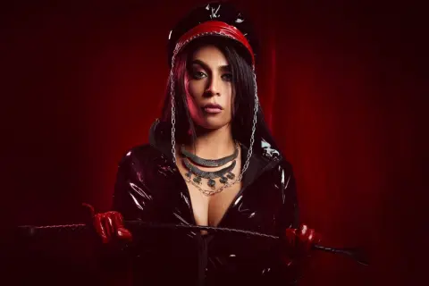 Mysterious woman in black latex outfit and red hat holding a whip against a dark red background.