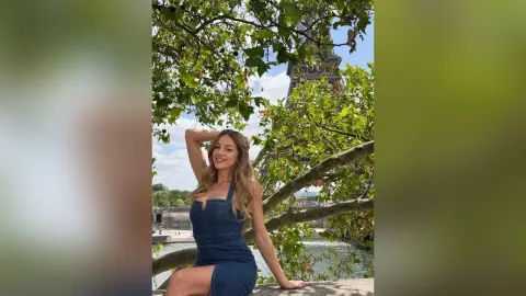 Smiling blonde woman in a denim dress relaxing under a tree with the Eiffel Tower in the background.