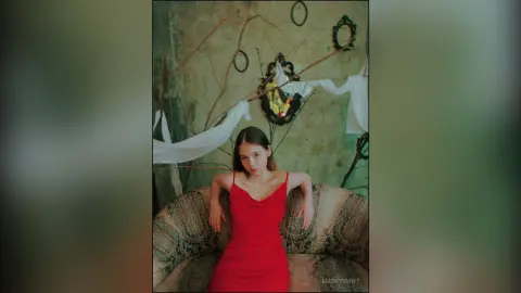 EmorySwift wearing a red slip dress, sitting on a patterned couch against a textured wall with decorative mirrors and branches.