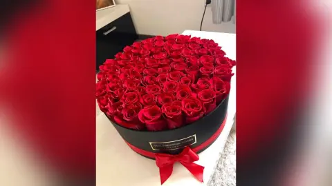 A large, round black gift box filled with a dense arrangement of bright red roses, decorated with a red ribbon and bow.