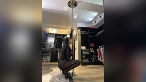NicoleBella poses naked against a silver dance pole in a dark bedroom setting, wearing sheer black stockings and high heels.