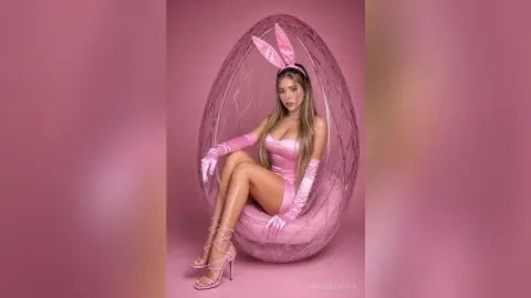 Glamorous woman in a pink bunny costume sitting in a decorative egg chair against a pink background.