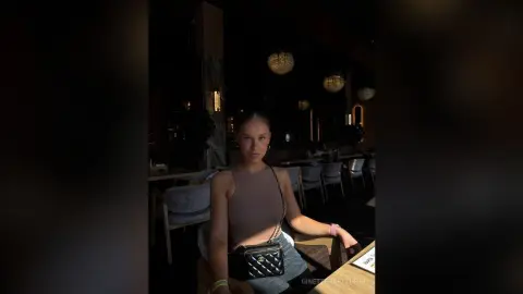 Young woman in a beige crop top and jeans sitting in a moody, dimly lit upscale restaurant.