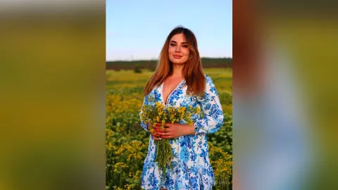 Smiling woman in a blue floral dress holding yellow wildflowers in a sunny summer meadow.