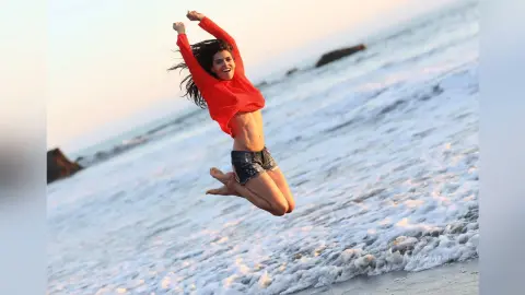 Joyful woman in a red sweater and denim shorts jumping on a sunny beach with ocean waves.