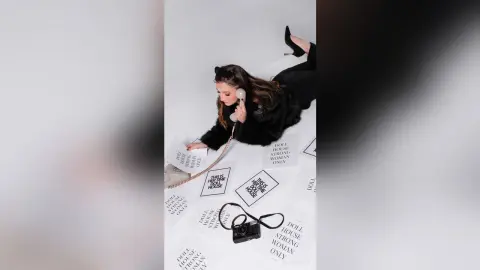 Fashionable woman in black outfit lying on floor with empowering 'Doll House Strong Woman Only' printed papers.