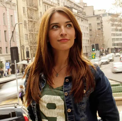 Smiling young woman in a denim jacket and green graphic tee standing on a sunny urban city street.