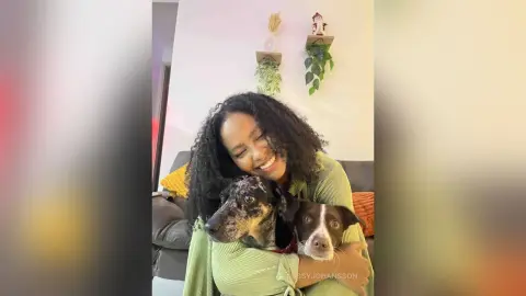 Smiling woman in a green ribbed sweater hugging a spotted dog on a cozy living room sofa.
