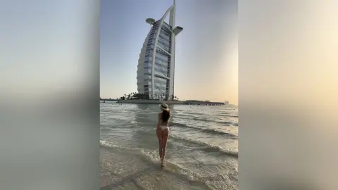 Woman in white bikini standing in the ocean with Burj Al Arab skyscraper in the background at sunset.