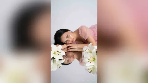 Elegant woman in a pink blouse gazing softly at white lilies in a dreamy, minimalist portrait.