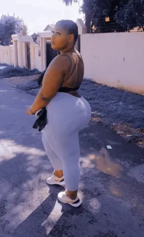 Curvy woman in grey leggings and black sports bra posing on a sunny residential street.