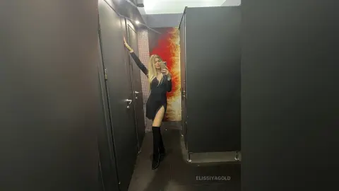 Glamorous blonde woman in a chic black mini dress and tall boots posing in a modern restroom mirror.