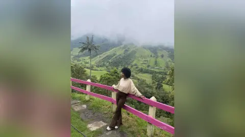 Woman in beige sweater and brown trousers leaning on a pink fence overlooking a lush green mountain valley.