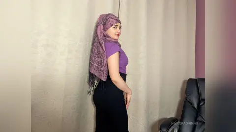 Elegant woman in a purple top and patterned mauve hijab posing confidently against a neutral curtain background.