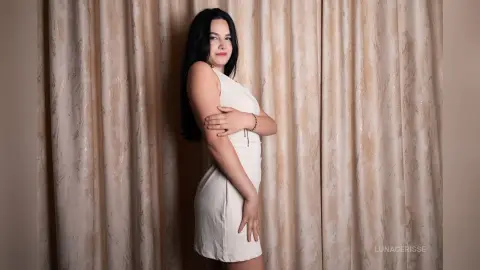 Young woman in a white sleeveless dress posing elegantly against beige textured curtains.