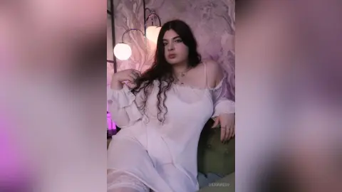 Young woman in a white flowing blouse sitting in a dreamy, soft-lit room with aesthetic lighting.