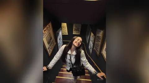 Smiling young woman in a white shirt and black crossbody bag posing in a dark hallway with vintage posters.