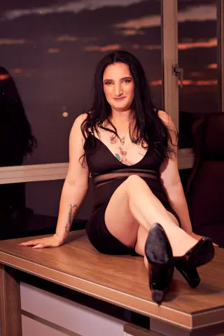 Smiling woman in a black cocktail dress sitting on a desk against a sunset window background.