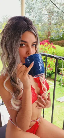 Young woman with wavy blonde hair in a red bikini sipping coffee on a sunny outdoor balcony.