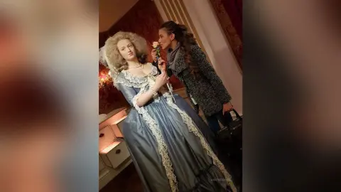 MontseFerrera wears a white lace and blue satin ballgown while holding a single rose to her lips.