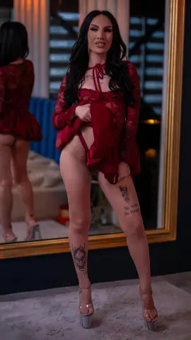 Woman in a red lace bodysuit standing in front of a mirror in a moody, stylish bedroom setting.