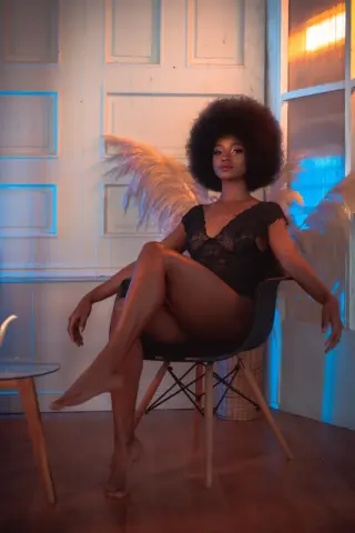 Elegant woman with afro hair and angel wings in black lace lingerie sitting on a chair in a dreamy blue room.