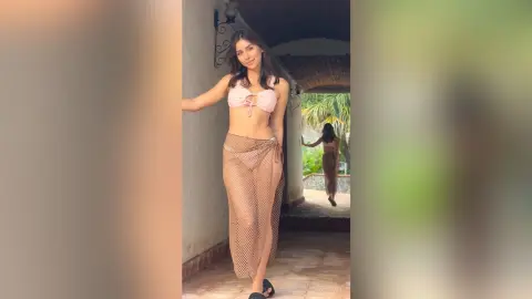 Smiling woman in a pink bikini top and mesh skirt posing in a sunlit tropical villa hallway.