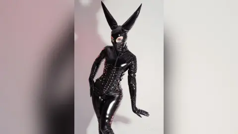 Woman in a black latex bunny bodysuit and mask posing in a studio with a moody pink lighting effect.
