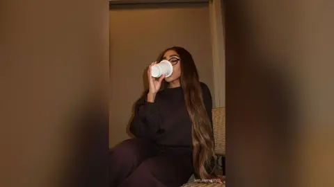 Young woman in a dark sweatshirt sipping coffee in a cozy, dimly lit indoor setting.