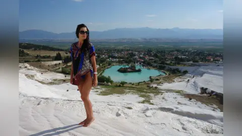 Woman in a patterned blue cover-up standing on white travertine terraces overlooking a scenic lake in Pamukkale.