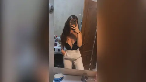 Young woman in a black bodysuit and white trousers taking a mirror selfie in a modern bathroom.