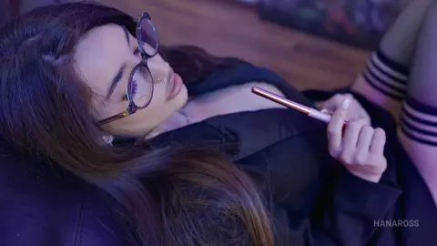 Young woman with glasses relaxing on a sofa in a dark room, holding a smartphone with a moody atmosphere.