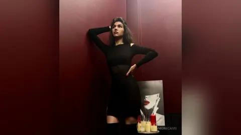 Stylish woman in a black long-sleeve outfit posing against a deep red wall in a moody, cinematic setting.
