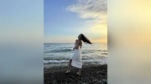 ChloeTeles stands on a dark sand beach at sunset, wearing a white backless dress with her long dark hair blowing in the wind.