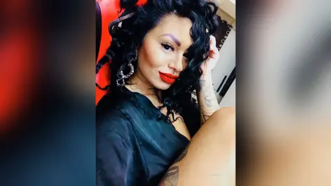 DeeaAnderson poses with long wavy black hair and bright red lipstick, wearing a black silk top and large silver hoop earrings.