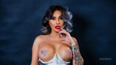 Glamorous woman with wavy hair and red lipstick wearing silver glitter heart pasties against a dark backdrop.
