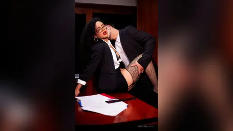 Glamorous woman in a black blazer and fishnet tights leaning back on a desk in a moody office setting.