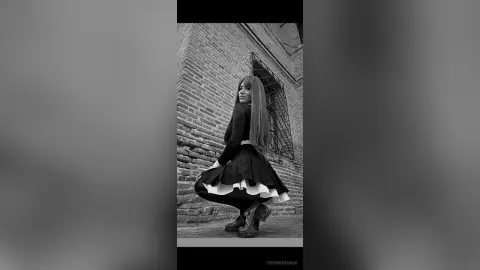 TiffanyAddams poses in a black long-sleeve top and a flared black and white skirt with dark boots against a brick wall.