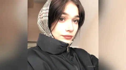 Young woman with dark hair wearing a plaid hoodie and black puffer jacket in a cozy indoor setting.