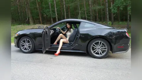 Stylish woman in a white top and red heels sitting in a black coupe parked on a forest road.