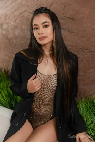 Elegant woman in a black blazer and sparkly mesh bodysuit posing against a textured stone wall.